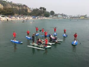 volunteers on standard paddle boards surrounding a wheel chair adapted stand up paddle boards. Everyone's arms up in celebration