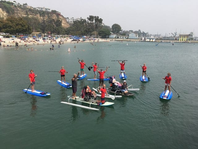 volunteers on standard paddle boards surrounding a wheel chair adapted stand up paddle boards. Everyone's arms up in celebration